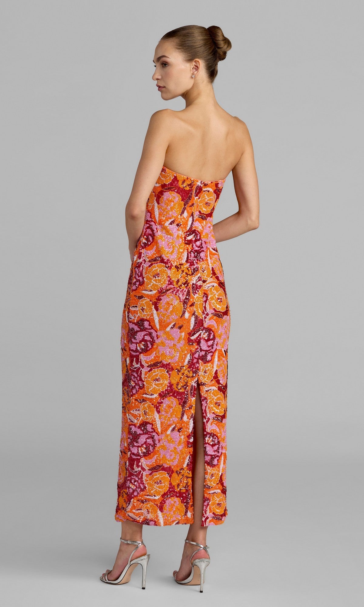 A woman stands in profile against a plain gray background, wearing the Mac Duggal 56385 Tea-Length Party Dressโa strapless design adorned with vibrant orange and pink florals, featuring a back slitโand paired with sparkling silver high-heeled sandals.